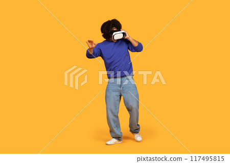 A man with a dark complexion wearing a blue sweatshirt and jeans stands against a yellow background. He is wearing a virtual reality headset and reacting to the immersive experience 117495815