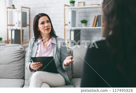 A woman therapist, dressed in a gray blazer and white pants, sits on a couch in an office setting. She is talking to her patient, who is sitting in front of her. Therapist has a clipboard in her hands A woman therapist, dressed in a gray blazer and white pants, sits on a couch in an office setting. She is talking to her patient, who is sitting in front of her. Therapist has a clipboard in her hands 117495828