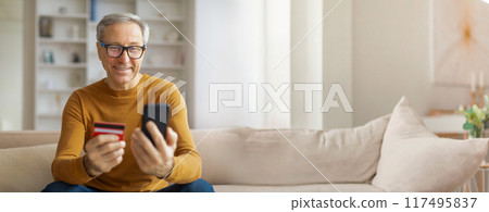 An older man is sitting on a sofa in his home, holding a smartphone in one hand and a credit card in the other. He is smiling and looking directly at the camera, copy space An older man is sitting on a sofa in his home, holding a smartphone in one hand and a credit card in the other. He is smiling and looking directly at the camera, copy space 117495837