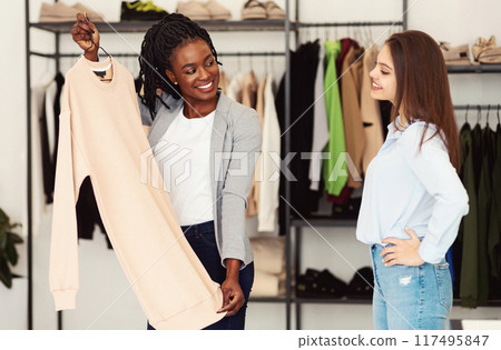 Two multiracial women of varying ages are standing in a clothing store, browsing through racks of clothes. They appear to be engaged in conversation as they inspect the different items on display. Two multiracial women of varying ages are standing in a clothing store, browsing through racks of clothes. They appear to be engaged in conversation as they inspect the different items on display. 117495847