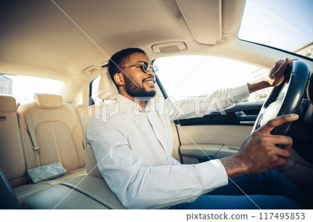African American man with a beard and sunglasses drives a car while wearing a white shirt. He appears to be enjoying the drive, looking out the window and smiling. 117495853