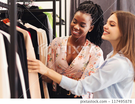 Two multiracial ladies are standing in front of a clothing rack indoors. They are browsing through the various garments, examining and comparing them 117495878