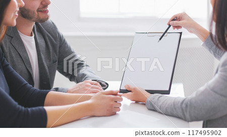 A businesswoman in a gray blazer is presenting a contract to a young couple in an office setting. She is holding a clipboard and pointing at the document with a pen. A businesswoman in a gray blazer is presenting a contract to a young couple in an office setting. She is holding a clipboard and pointing at the document with a pen. 117495902