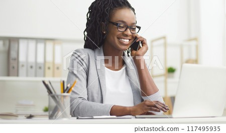 Cheerful young African American lady manager in formal wear, using a laptop at her office desk, speaking on the phone. Cheerful young African American lady manager in formal wear, using a laptop at her office desk, speaking on the phone. 117495935
