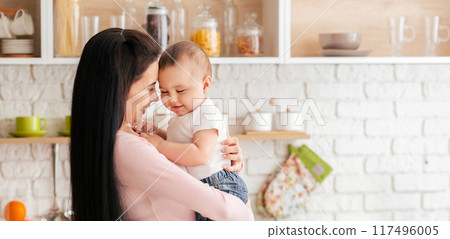 A woman is holding a baby in her arms. The woman is standing and cradling the baby securely, with a gentle expression on her face. Both individuals are dressed in casual clothing. 117496005