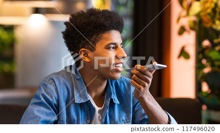 A young black guy is sitting in a modern cafe, holding his smartphone and using a voice assistant to perform tasks or send messages. 117496020