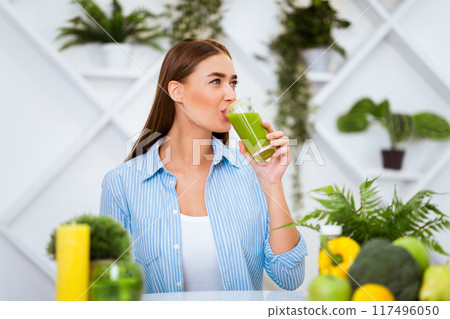 A woman in a blue shirt sits at a table in a kitchen and drinks a green smoothie. Surrounding her are fruits and vegetables, and there are plants in the background. A woman in a blue shirt sits at a table in a kitchen and drinks a green smoothie. Surrounding her are fruits and vegetables, and there are plants in the background. 117496050