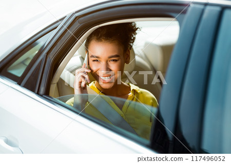 A young black woman with dark hair and a yellow shirt is smiling as she talks on her phone. She is sitting in the back seat of a white car, with the window open. A young black woman with dark hair and a yellow shirt is smiling as she talks on her phone. She is sitting in the back seat of a white car, with the window open. 117496052