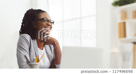 Beaming African American businesswoman working on her laptop at her desk in a trendy office environment, looking at copy space 117496098