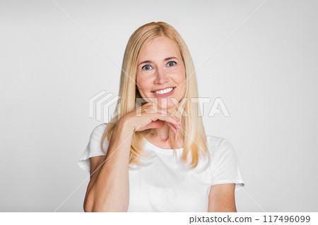 A portrait of a woman with long, blond hair smiling gently at the camera. She is wearing a white t-shirt and is resting her chin on her hand. The background is a simple, light gray. A portrait of a woman with long, blond hair smiling gently at the camera. She is wearing a white t-shirt and is resting her chin on her hand. The background is a simple, light gray. 117496099