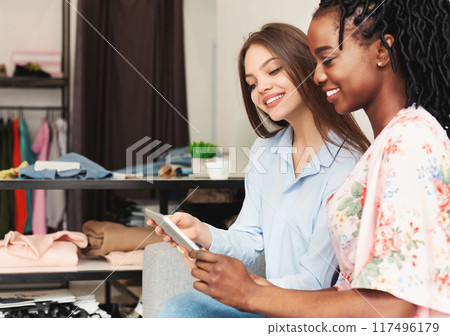 Two multiethnic women are seated on a couch, both looking intently at digital tablet held between them, visiting fashion store, checking new collection Two multiethnic women are seated on a couch, both looking intently at digital tablet held between them, visiting fashion store, checking new collection 117496179