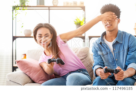 African American girl is covering her boyfriends eyes with her hand while he plays video games on the couch. They are both smiling and having fun. They are likely in a living room or home setting. 117496185