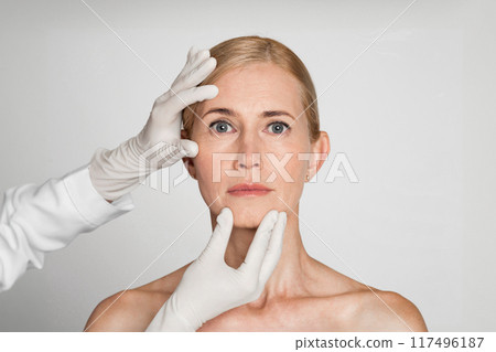 A woman with blonde hair and a serious expression is receiving a facial examination. Two hands, wearing white gloves, are touching her forehead and chin. 117496187