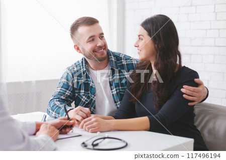 A young couple sits at a table in a doctors office, engaged in conversation with the doctor off-camera. The man is wearing a plaid shirt and the woman is wearing a black long-sleeve shirt. A young couple sits at a table in a doctors office, engaged in conversation with the doctor off-camera. The man is wearing a plaid shirt and the woman is wearing a black long-sleeve shirt. 117496194