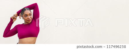 Sexy athletic young black woman in pink sportswear posing on white blank wall background, looking at copy space for advertisement, sportswoman demonstrating perfect body curves, using earpods 117496238