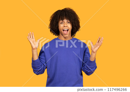 Young man with curly hair wearing a blue sweatshirt and standing in front of a yellow background. He has a surprised expression on his face and is holding both of his hands up in the air. Young man with curly hair wearing a blue sweatshirt and standing in front of a yellow background. He has a surprised expression on his face and is holding both of his hands up in the air. 117496266