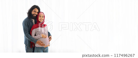 A happy Muslim couple is shown inside their home. The man, with a beard, has his arm around his pregnant wife, who is wearing a hijab. They are smiling at the camera, copy space 117496267