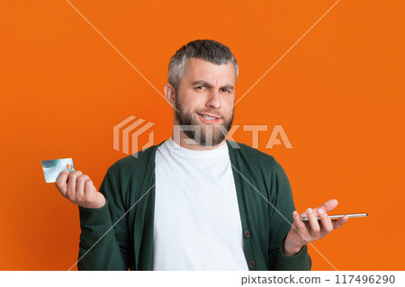A bearded man stands against an orange background, holding a credit card in one hand and a smartphone in the other. He looks puzzled. A bearded man stands against an orange background, holding a credit card in one hand and a smartphone in the other. He looks puzzled. 117496290