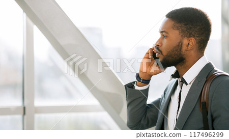 A professional black man, talking on the phone at the entrance of an airport. The environment is bustling and contemporary, emphasizing the business travel experience. 117496299