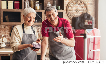 Senior couple food vloggers cooking together at home kitchen, recording video on camera, showing organic vegetables 117496310