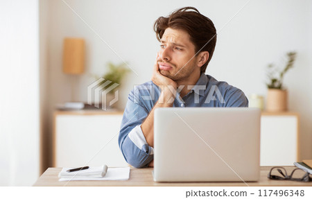 A pensive young man gazes out the window at his desk, chin on hand, with a laptop in front of him and a notepad and pen nearby. 117496348