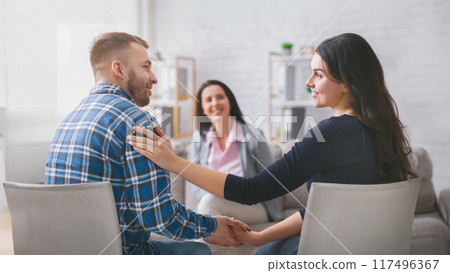 A couple sits facing each other in a therapy session, holding hands. The woman is smiling at her partner, while the man looks happy and relaxed. A lady counselor sits behind them A couple sits facing each other in a therapy session, holding hands. The woman is smiling at her partner, while the man looks happy and relaxed. A lady counselor sits behind them 117496367