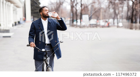 An executive black guy is cycling to the office through downtown, drinking water. The mood is lively and productive, with an urban backdrop highlighting a modern and active commute. An executive black guy is cycling to the office through downtown, drinking water. The mood is lively and productive, with an urban backdrop highlighting a modern and active commute. 117496372