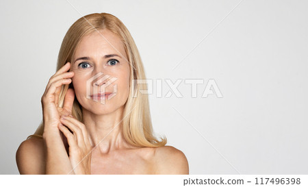 A portrait of a middle-aged blonde woman touching her face with one hand. She is wearing no makeup and has a natural expression. She is standing against a white backdrop, copy space 117496398