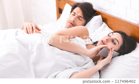 A woman lies in bed, talking on her phone while her partner sleeps soundly beside her. The woman expression suggests she is engaged in a private conversation. 117496402