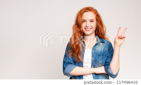 A woman with vibrant red hair is standing, raising her hand to form the peace sign gesture, symbolizing harmony and unity. She is wearing casual attire and appears confident and expressive. A woman with vibrant red hair is standing, raising her hand to form the peace sign gesture, symbolizing harmony and unity. She is wearing casual attire and appears confident and expressive. 117496410