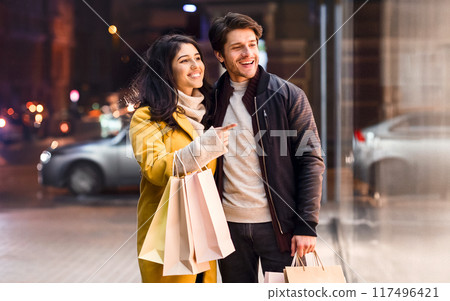 A man and woman are standing on a bustling city street, holding multiple shopping bags. They appear to have been on a shopping spree. 117496421