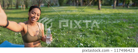 Cheery young black lady vlogger taking selfie or speaking to followers remotely on mobile phone, doing yoga or meditation at city park. Sports blogger, online coach and distance training, copy space 117496423