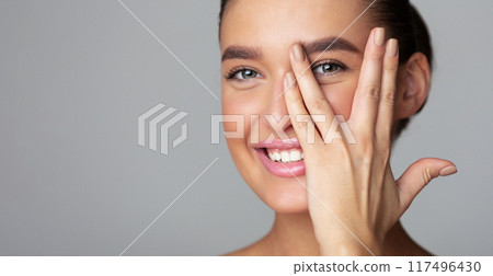 Cheerful young woman covering her face with palm, peeking from fingers and smiling, grey background, panorama with copy space 117496430