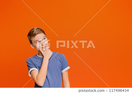Pensive school boy thinking about something, looking up at free space, orange studio background 117496434