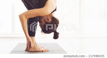 A woman is seen practicing yoga indoors, performing a forward fold pose. The setting is brightly lit with natural light streaming in from large windows, panorama with copy space 117496435