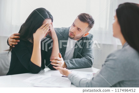 The image shows a man comforting a woman who is upset and covering her face with her hands. They are sitting at a table with a counselor, likely discussing a difficult situation. 117496452
