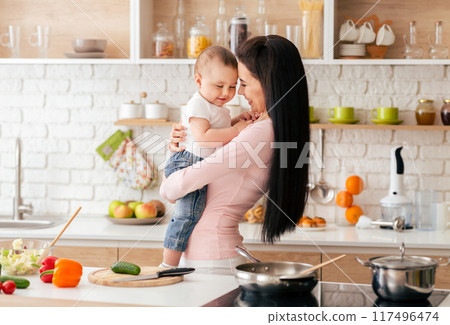 A woman standing in a kitchen, holding a baby in her arms. She appears to be looking down at the baby with a gentle expression. 117496474