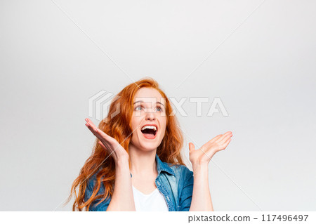 A woman with bright red hair and a denim jacket is looking upwards with an open-mouthed smile, her arms raised in a cheerful and surprised gesture, copy space A woman with bright red hair and a denim jacket is looking upwards with an open-mouthed smile, her arms raised in a cheerful and surprised gesture, copy space 117496497