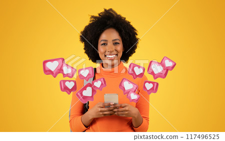 Smiling millennial African American woman with curly hair, wearing an orange turtleneck, texts boyfriend on her phone while carrying a grey backpack on a vibrant yellow background, studio 117496525