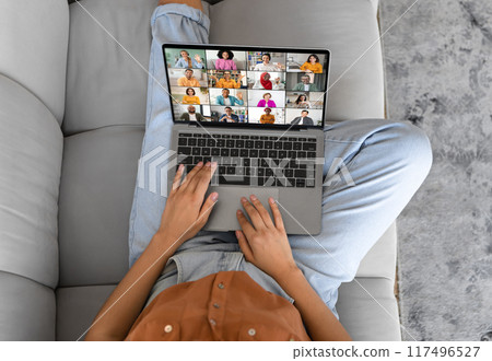 Unrecognizable lady is sitting on a couch and using a laptop to video call with several other people. The person is using the trackpad on the laptop to navigate the call. 117496527