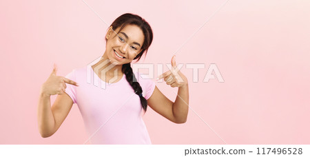 A cheerful, young African American girl with a braid is smiling brightly and pointing at herself while standing against a light pink background during the daytime. 117496528