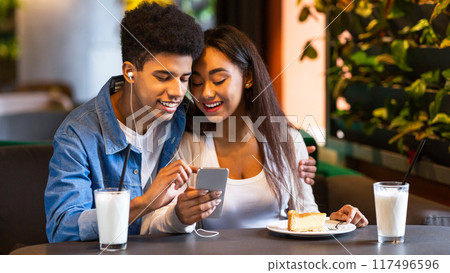 Two African American teenagers are sitting at a cozy cafe, enjoying milkshakes and a slice of cake. They are smiling and watching something on a smartphone together. Two African American teenagers are sitting at a cozy cafe, enjoying milkshakes and a slice of cake. They are smiling and watching something on a smartphone together. 117496596