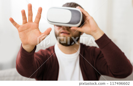 A bearded man is wearing a virtual reality headset, immersing himself in a digital environment. His facial features are partially obscured by the device as he interacts with the virtual world 117496602