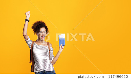 African American young woman with curly hair exudes excitement as she raises one fist in triumph. She is holding a passport and tickets on yellow background, copy space 117496620
