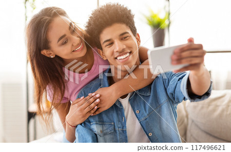 African American teen couple taking a selfie at home. The girl is standing behind the guy, with her arms wrapped around his shoulders. They both look happy and in love 117496621