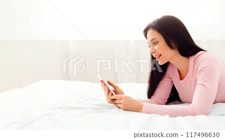 A woman is laying on a bed, focused on a cell phone screen in her hands. She appears engrossed in the device, with a relaxed posture. The room is dimly lit, emphasizing the screens glow. A woman is laying on a bed, focused on a cell phone screen in her hands. She appears engrossed in the device, with a relaxed posture. The room is dimly lit, emphasizing the screens glow. 117496630