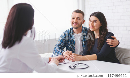 A young couple sits at a table with woman doctor, listening intently as she speaks. The woman is sitting slightly in front of her husband, with his arm around her shoulder A young couple sits at a table with woman doctor, listening intently as she speaks. The woman is sitting slightly in front of her husband, with his arm around her shoulder 117496631