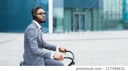 An African American businessman is cycling to the office through the city. The setting is contemporary and busy, emphasizing an active and health-conscious commute. An African American businessman is cycling to the office through the city. The setting is contemporary and busy, emphasizing an active and health-conscious commute. 117496632