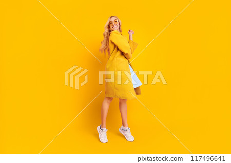 A happy young woman wearing a yellow coat and white sneakers, stands with a bright smile on her face and poses playfully in front of a yellow background. 117496641