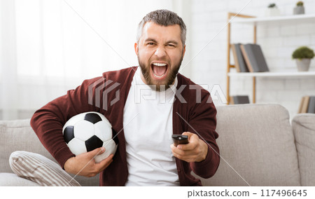 A man is shown holding a soccer ball in one hand and a remote control in the other. He appears to be multitasking or switching between watching TV and playing soccer. A man is shown holding a soccer ball in one hand and a remote control in the other. He appears to be multitasking or switching between watching TV and playing soccer. 117496645